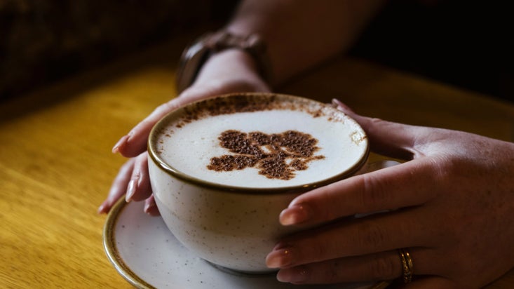 Image of a female caresses a hot cup of coffee with the National Trust oak leaf logo stencilled in chocolate on the top.